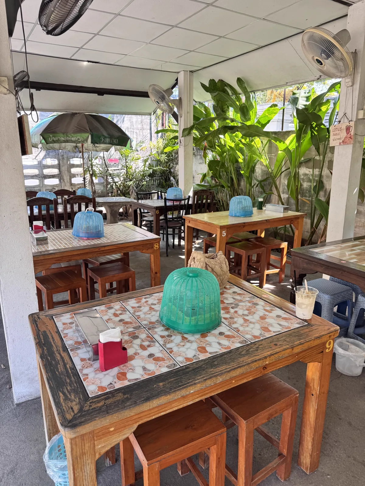 Open-air dining area with wooden tables, tiled tops, stools and potted banana plants