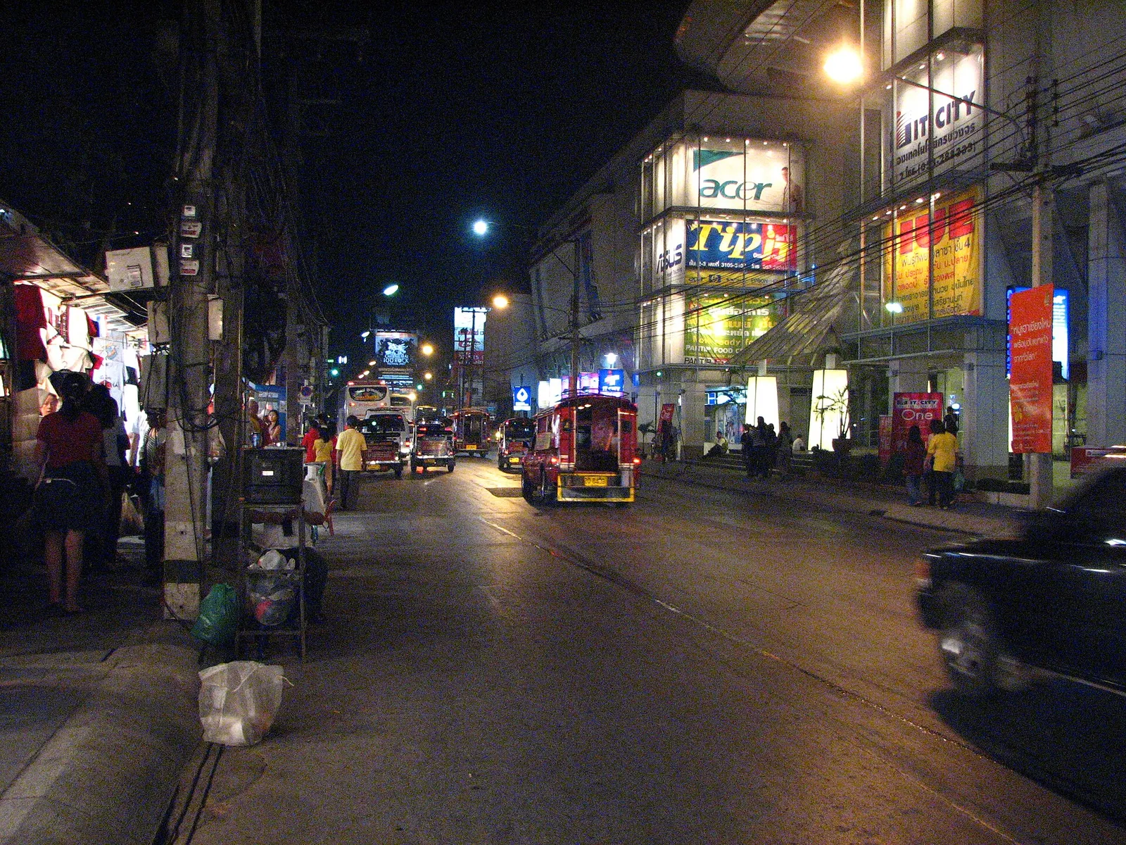 Busy night street lined with market stalls and shopfronts under street lights, with a red songthaew on the road