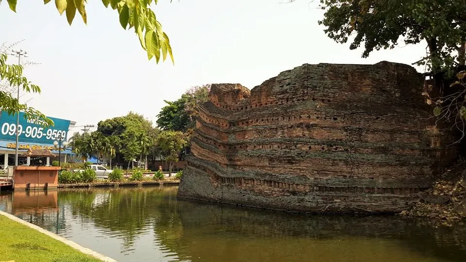 Weathered brick wall corner rising from a moat, with trees and a footpath beside the water